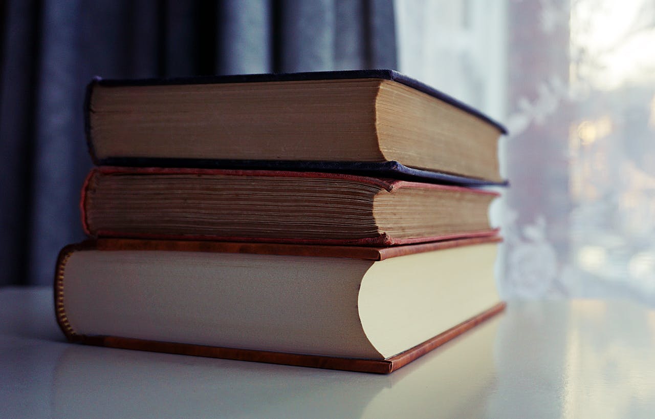 A stack of old books sits on a shelf by a sunlit window, capturing a sense of quiet study.